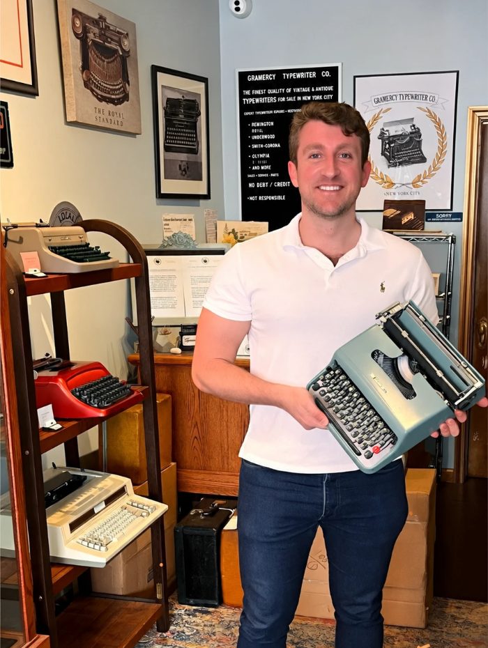 Wade in a typewriter shop in New York, smiling, holding a sage-green 1956 Olivetti Lettera 22 typewriter